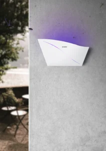 Real-life installation showing the medium size white GOKDA Nest 2 mosquito trap wall-mounted in a home backyard environment, with table and chairs behind.